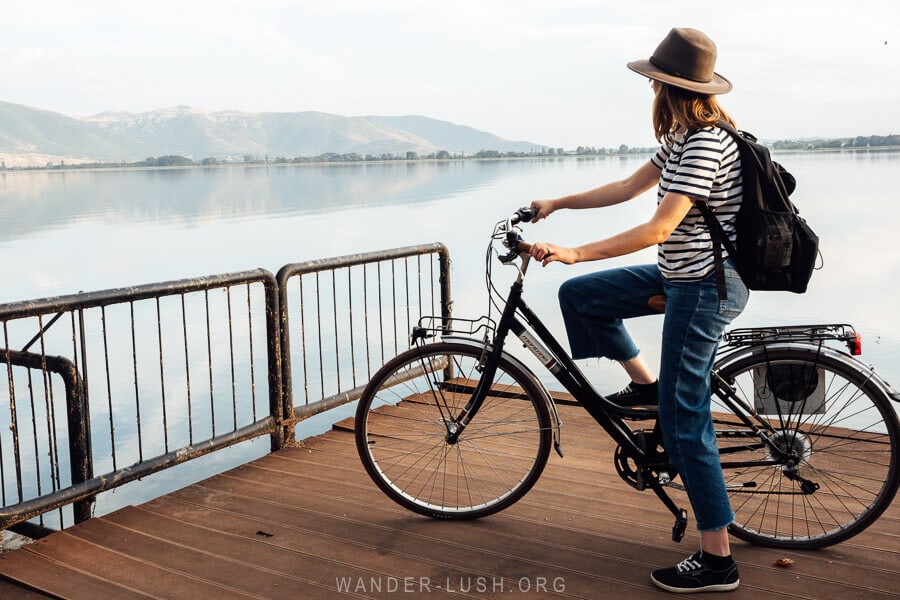 A woman in jeans riding a bicycle on a path along the edge of Lake Orestiada in Kastoria, Greece.