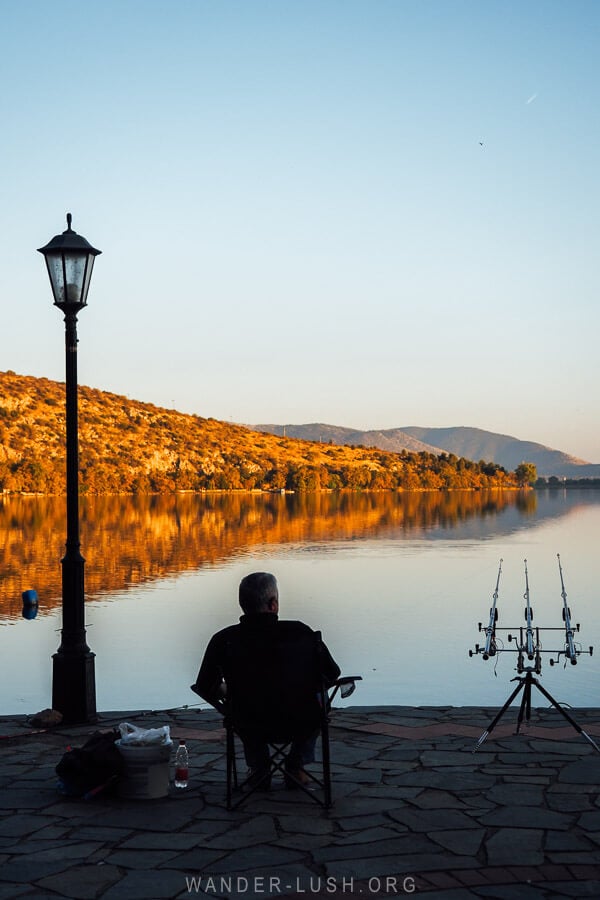 A man sitting on the edge of Lake Orestiada with three fishing rods at sunset.