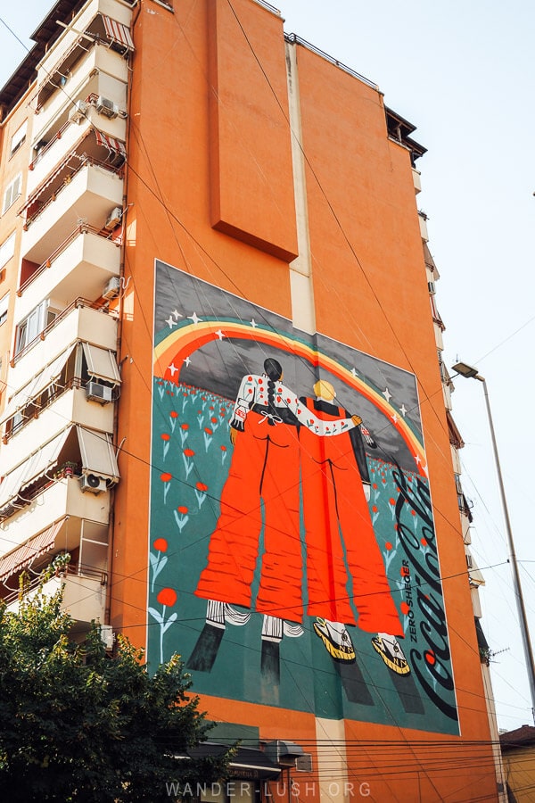 Street art on the side of an apartment building in Tirana shows two friends arm in arm under a rainbow.