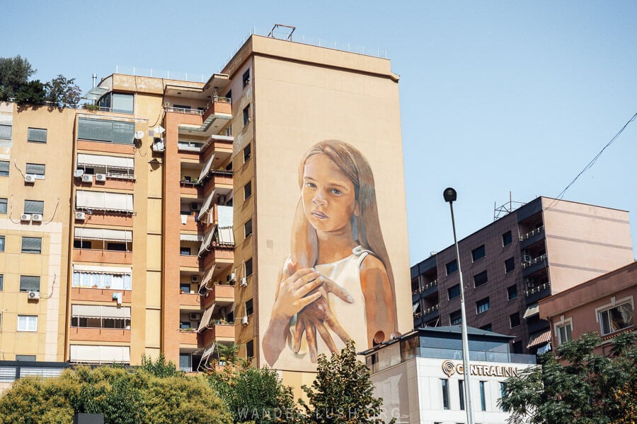 A street art mural in Tirana depicting a young girl with her mother's hand draped over her shoulder.