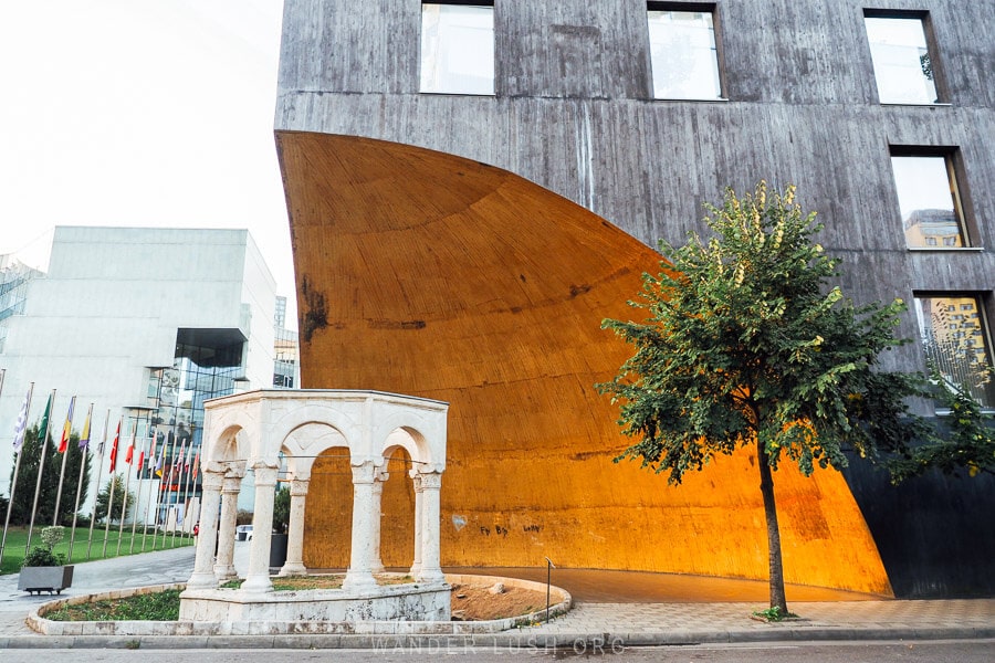 An ancient stone tomb on a street corner in Tirana Albania with a modern building arcing over it and a small tree nearby.