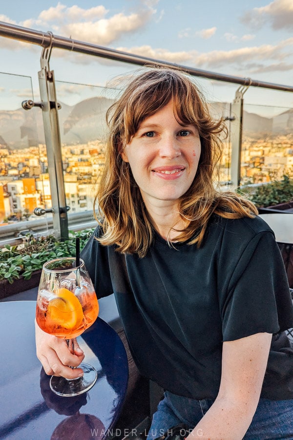 A woman dressed in black holding a cocktail at a sky bar in Tirana, Albania, with the city skyline and mountains in the distance.