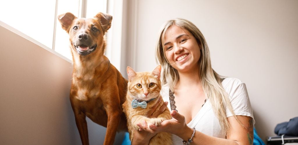 A woman sitter with a cat and a dog beside the window. 