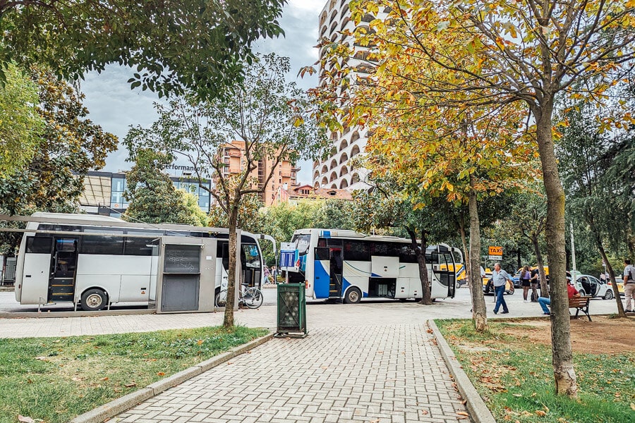 White buses wait on the edge of a park in Tirana to take tourists to the airport.