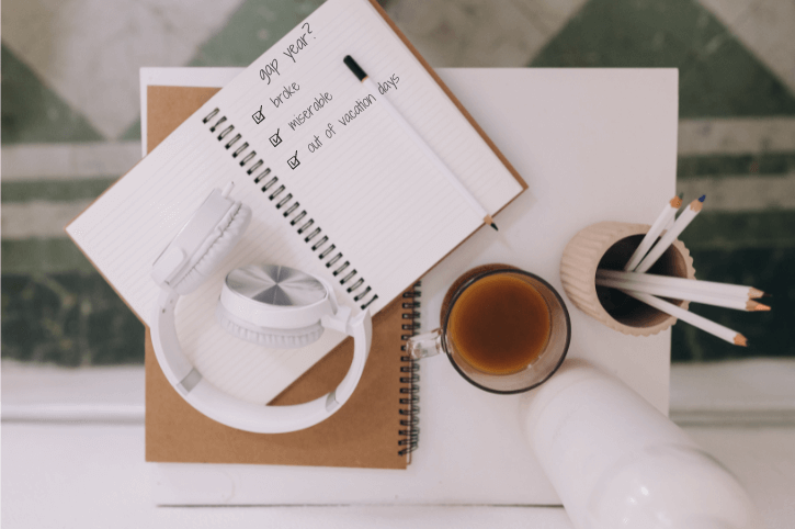 Flatlay of casual office setup with a notebook open that reads "gap year?" with checkboxes ticked that read "broke, miserable, and out of vacation days."