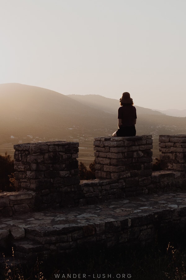 A woman sitting on the ruins of the stone Berat Castle at sunset.