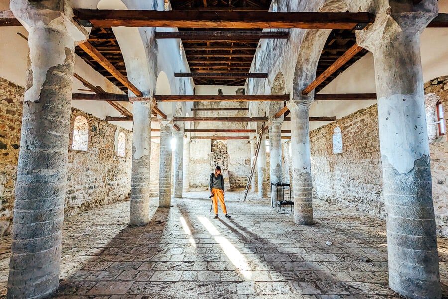 A woman dressed in yellow pants standing inside a stone church in Voskopoja, Albania, with the evening sun streaming in.
