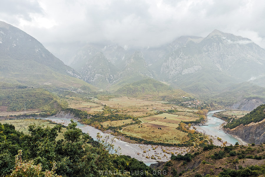 The Vjosa River Bend viewpoint in Albania, with a view of the river, fields, and distant limestone mountains.