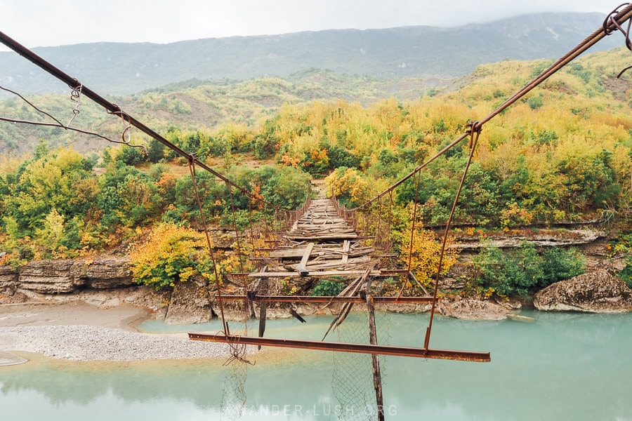 An abandoned suspension bridge in southern Albania stretches over a river with fall colours in the background.