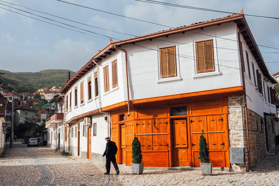 A man in a hat strides past a heritage building with wooden details in Pogradec, Albania.