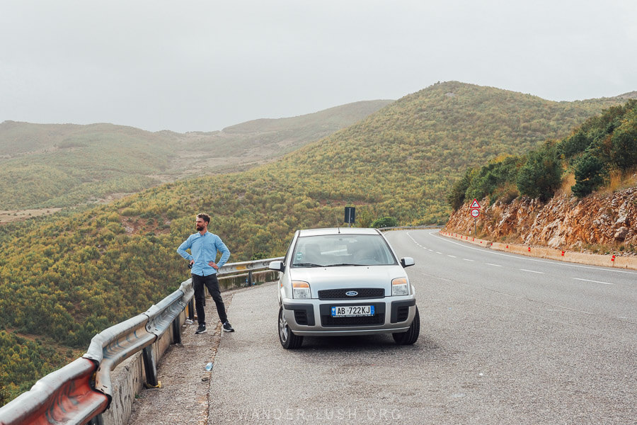 A man standing next to a silver car on a highway along Lake Ohrid in Albania.