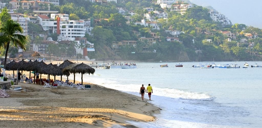 A couple is walking on the shore with a small pavilion on the side and some floating boats on the sea. and buildings in the background. 