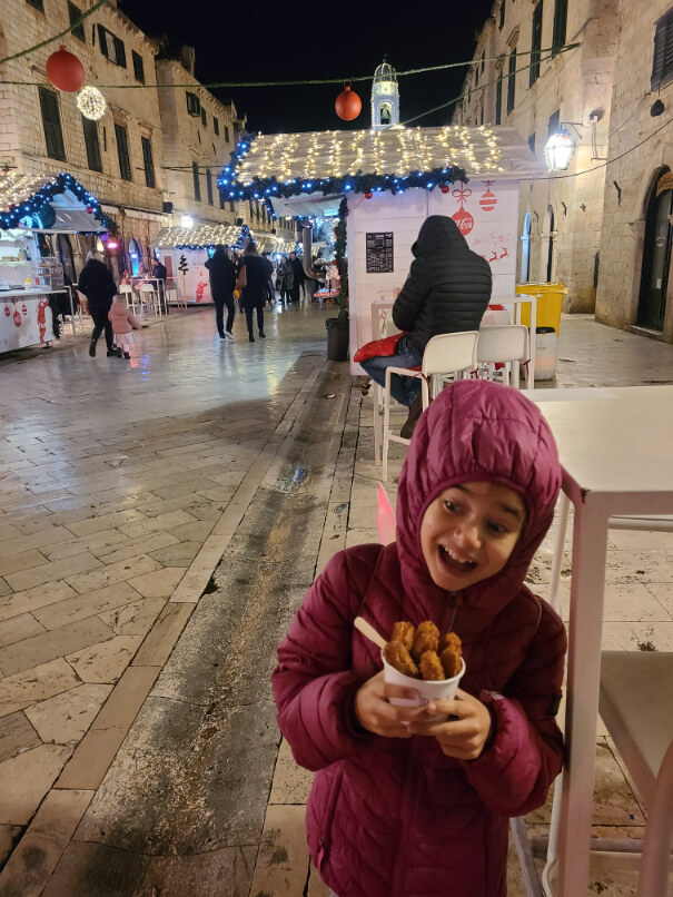 Little girl in a pink coat and hood looks excited over a cup of churros in front of the Dubrovnik Christmas Market