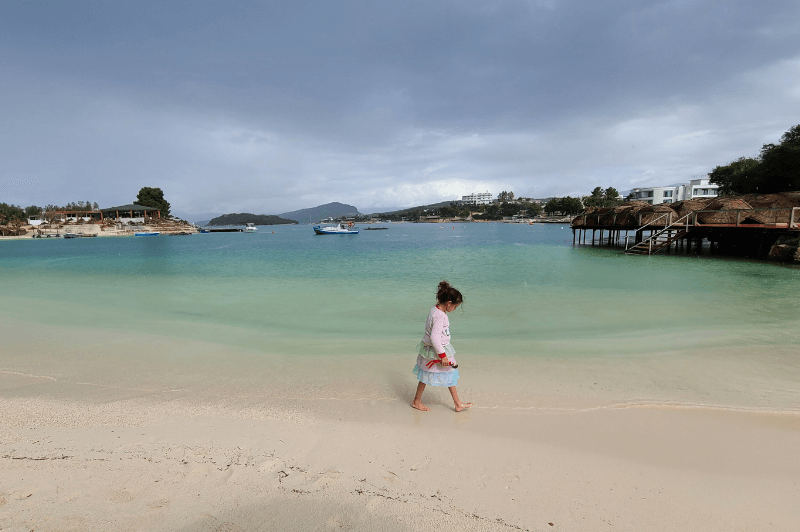 A small girl in a pink dress walks barefoot in the shallow sandy water of Lori Beach in Ksamil Albania