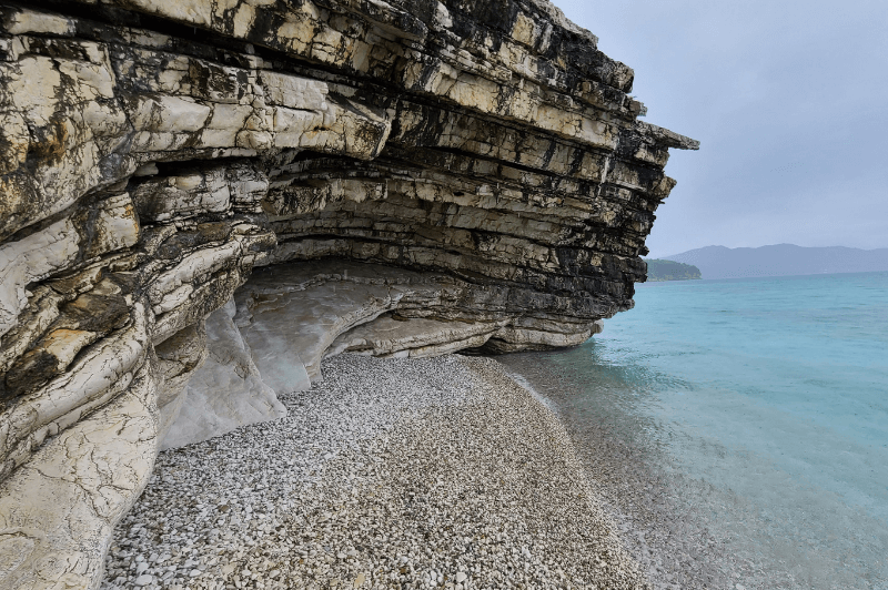 Round white pebbles line Mirror Beach Albania with a rock cliff overhanging the crystal blue waters