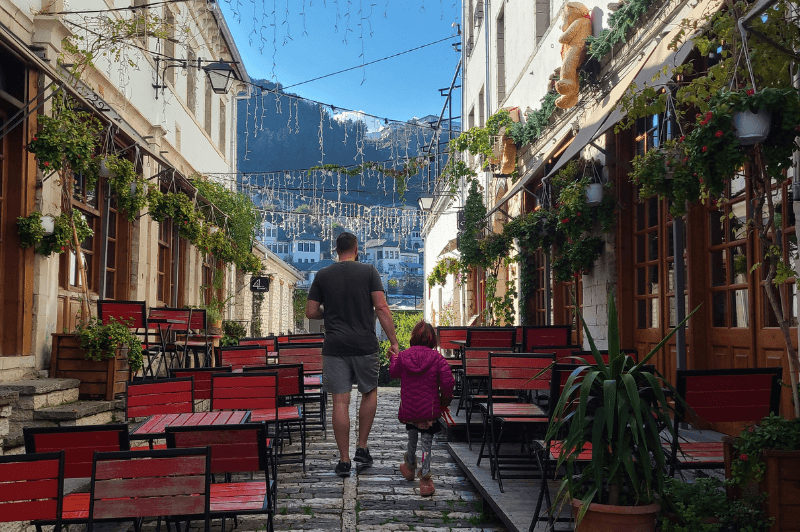 A man and his daughter walk through a narrow street in Gjirokaster Albania. The street is lined with hanging plants and red chairs.