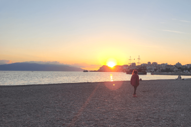 Sunset over saranda beach with a girl standing on the sand and buildings profiled in the background