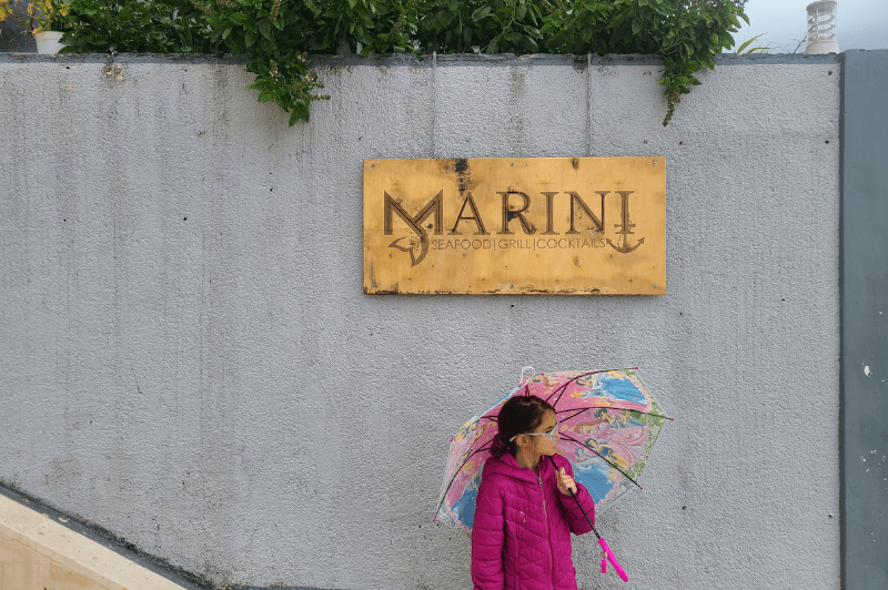 girl stands under the wood sign at Marini restaurant in Saranda holding an umbrella