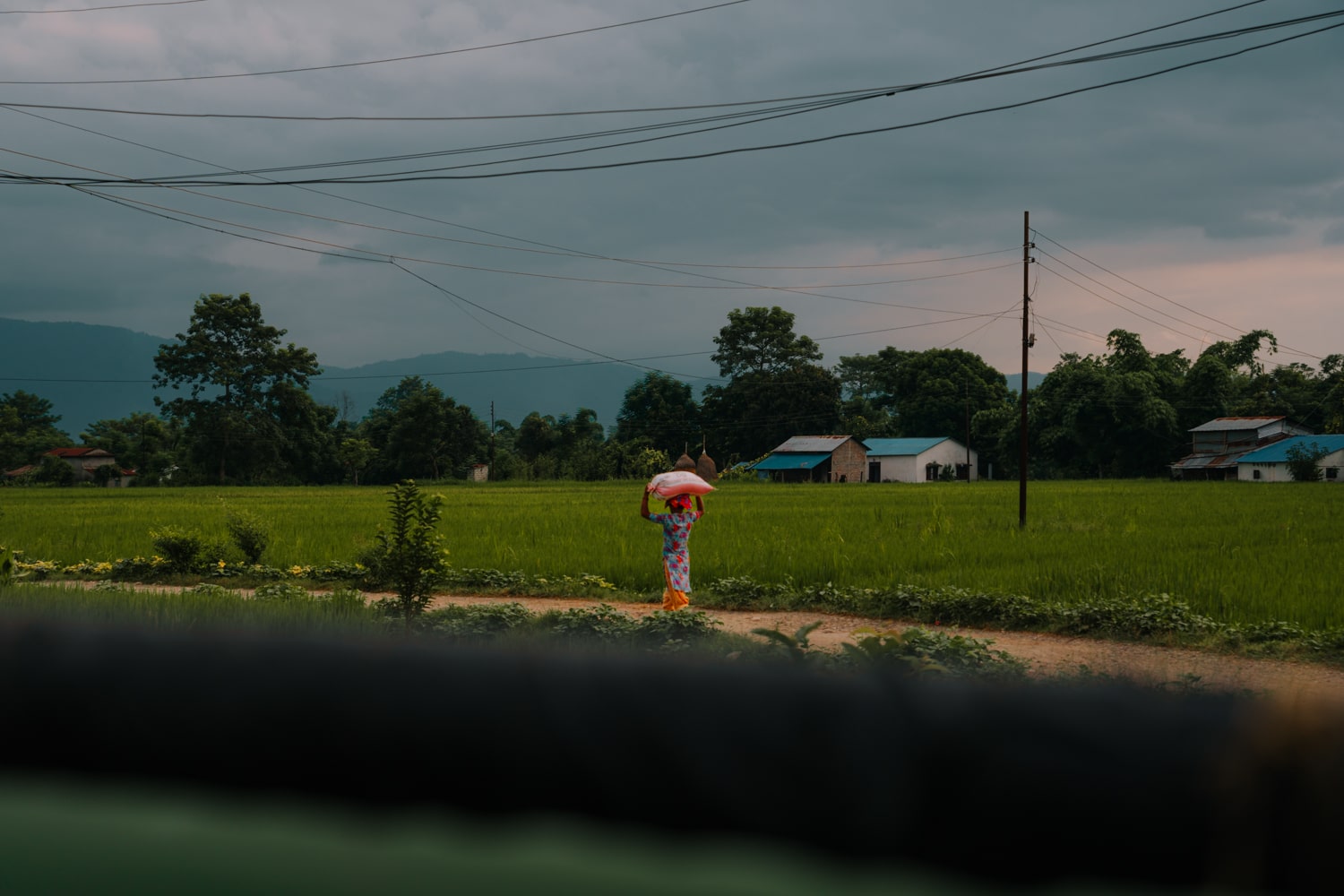 A local Tharu woman walking down dirt road in Barauli village carrying a bag of rice on her head.