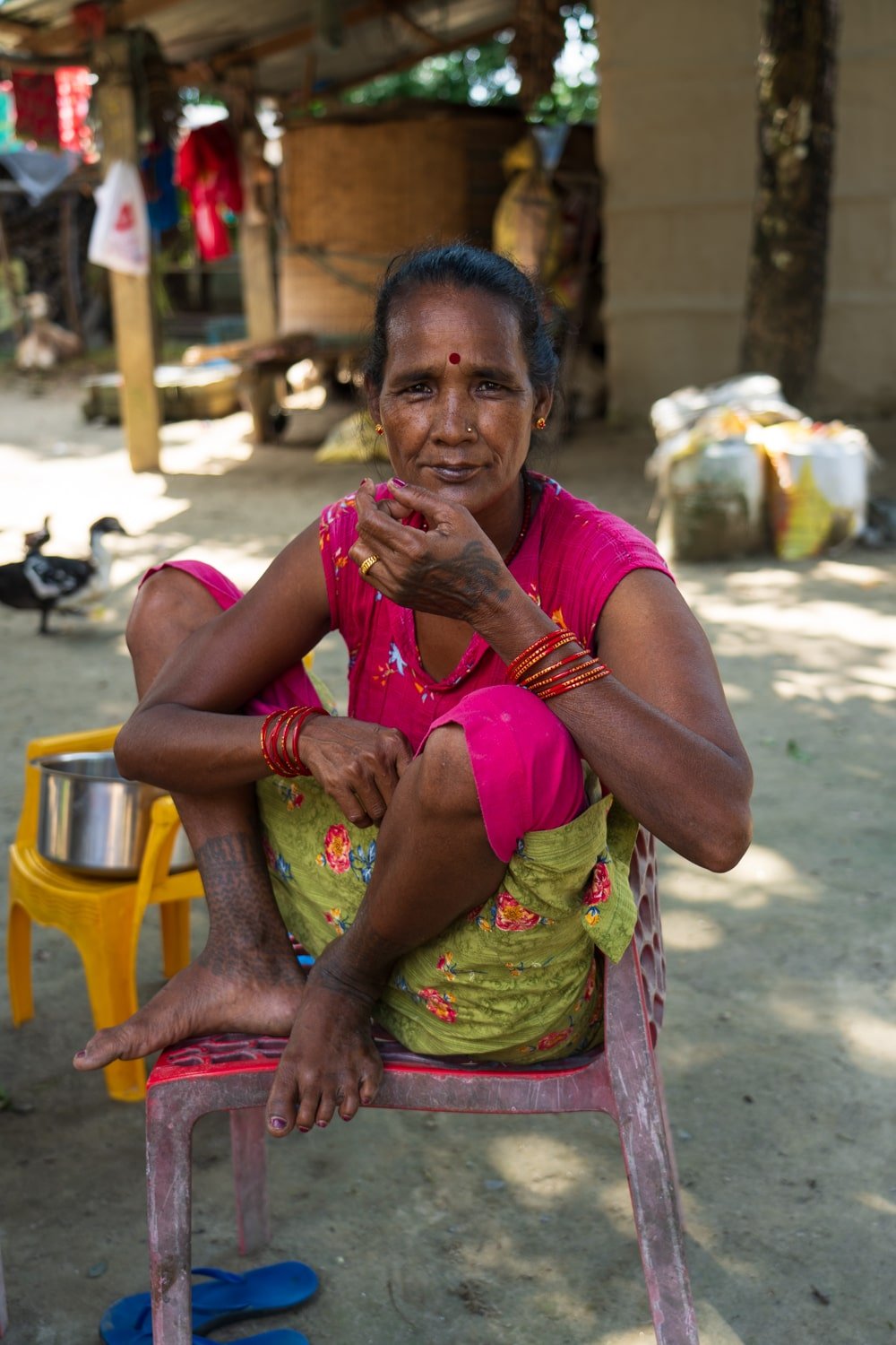 A local Tharu woman with ankle and hand tattoos squats on a chair at her home dressed in bright pink and green clothes.