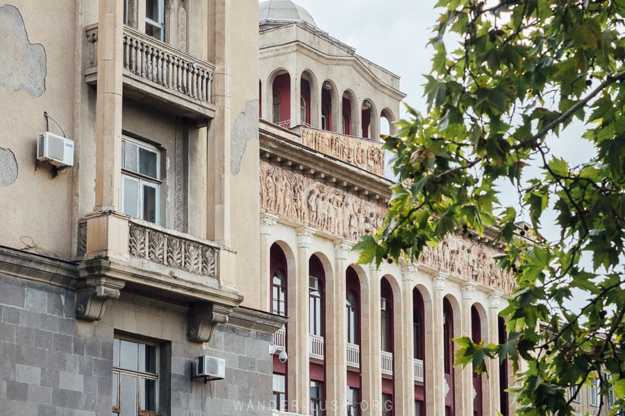 Side view of the Rustavi Metallurgy Plant facade, with bas-relief sculpture visible through the leaves of a pomegranate tree.