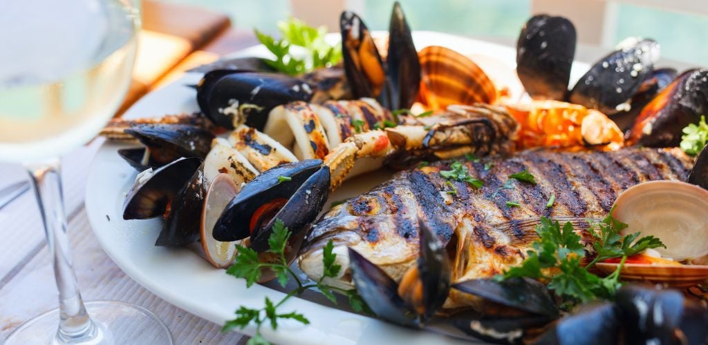 A grilled fish, clamp, squid, and mussels on the plate and a glass of wine. 