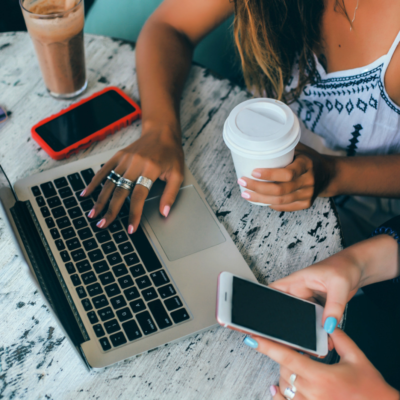 Two-Digital-Nomads-Looking-At-One-Laptop-And-Their-Mobile-Cell-Phones-With-A-Takeaway-Coffee