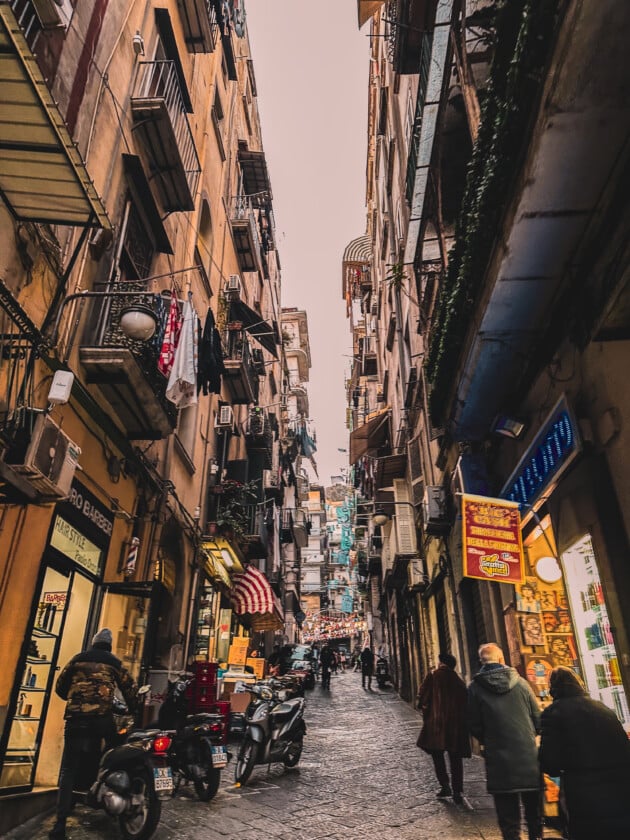 A bustling city street in the Spanish Quarter of Naples, Italy, flanked by colorful buildings and filled with vibrant activity.