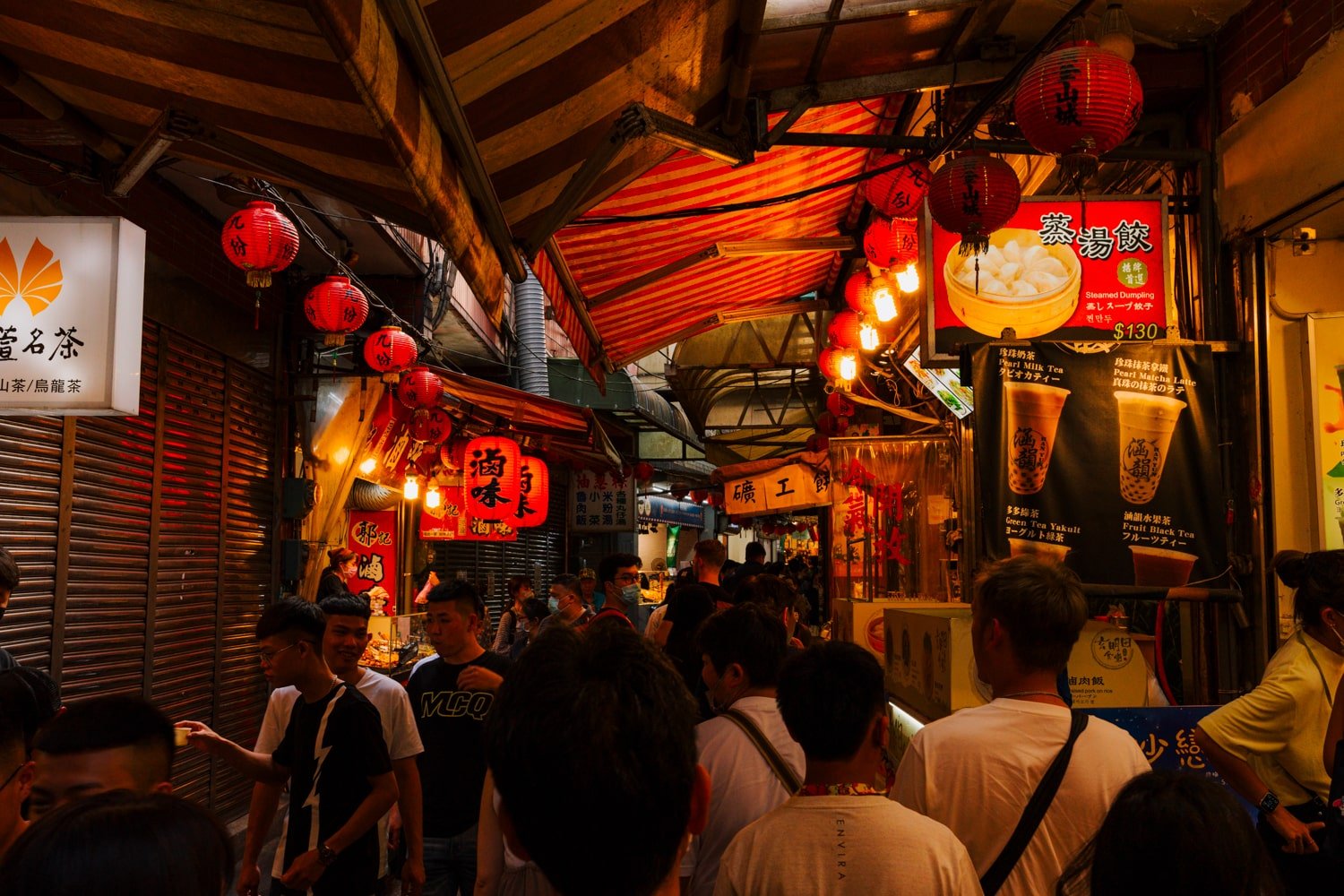 Tourists walk down crowded Jiufen Old Street with street food vendors and red lanterns.