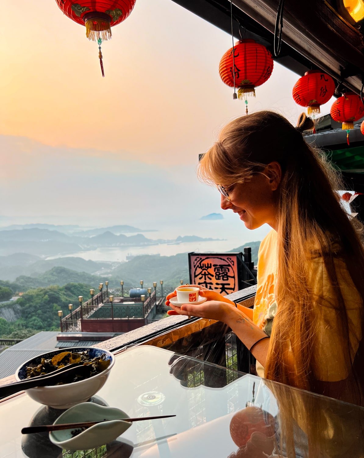 Female traveler holds cup of Taiwanese tea at Amei Teahouse overlooking Jiufen and Keelung Bay at sunset.