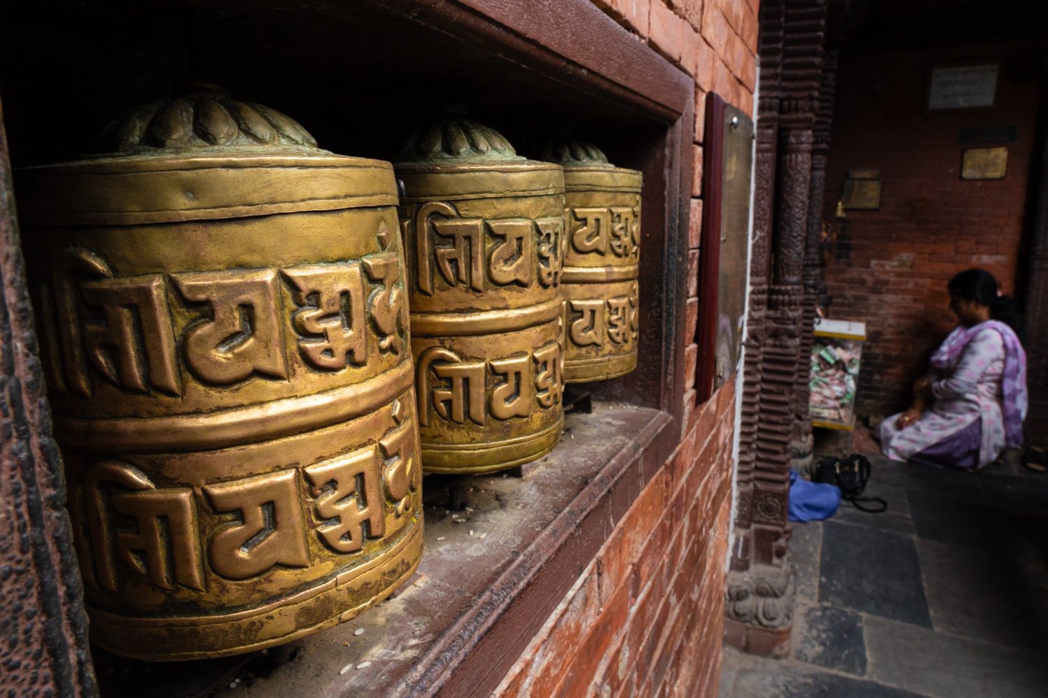 A woman prays in the corner beside a row of golden prayer wheels.