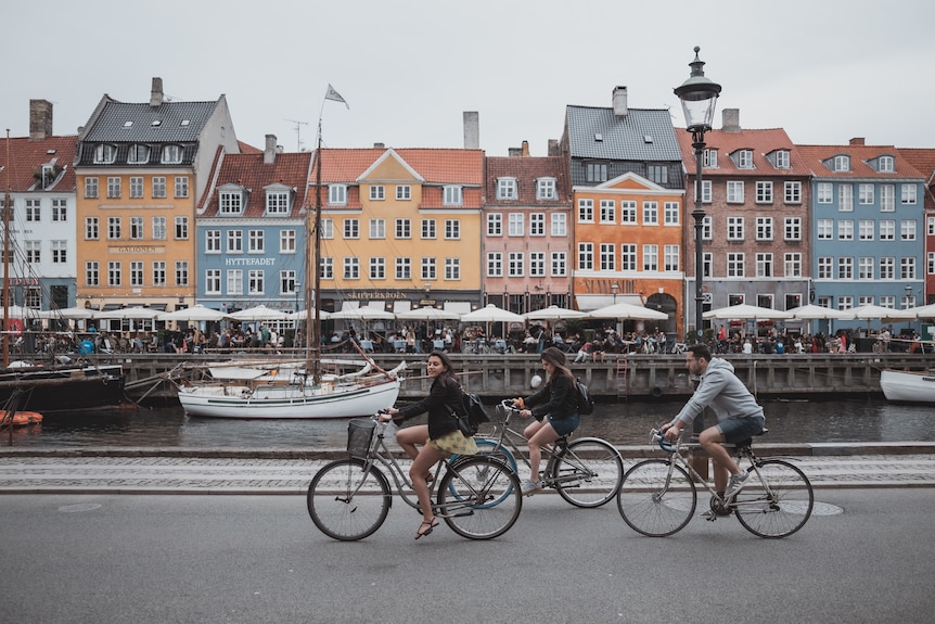 Three people ride bicycles alongside a lake with colourful pastel buildings in the background