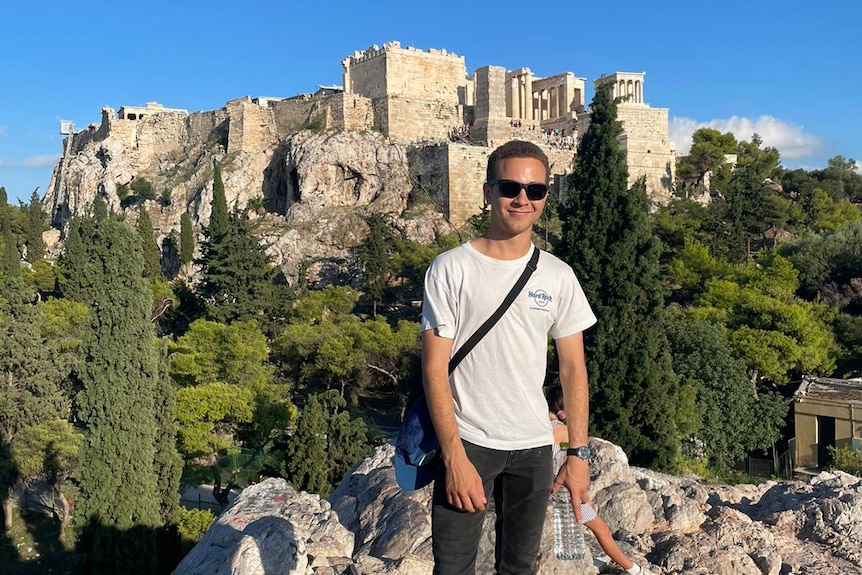 A man wearing a white t-shirt stands on a rocky cliff with trees behind him