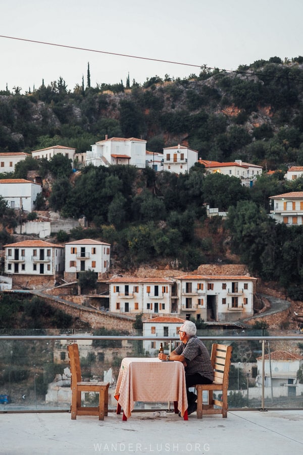 A man sitting at a wooden table in front of a viewpoint in Dhermi.