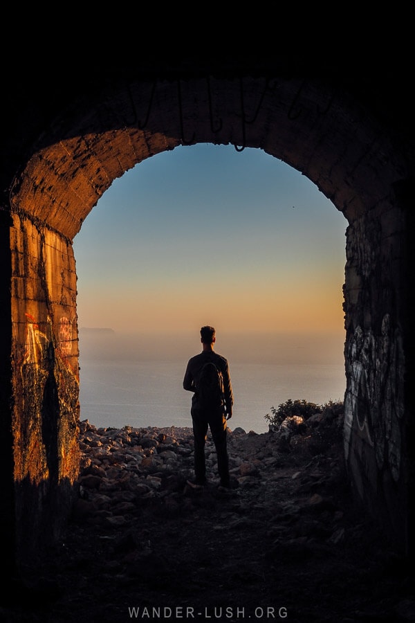 A man stands at the opening to a bunker in Albania, with a view of the sea at sunset.