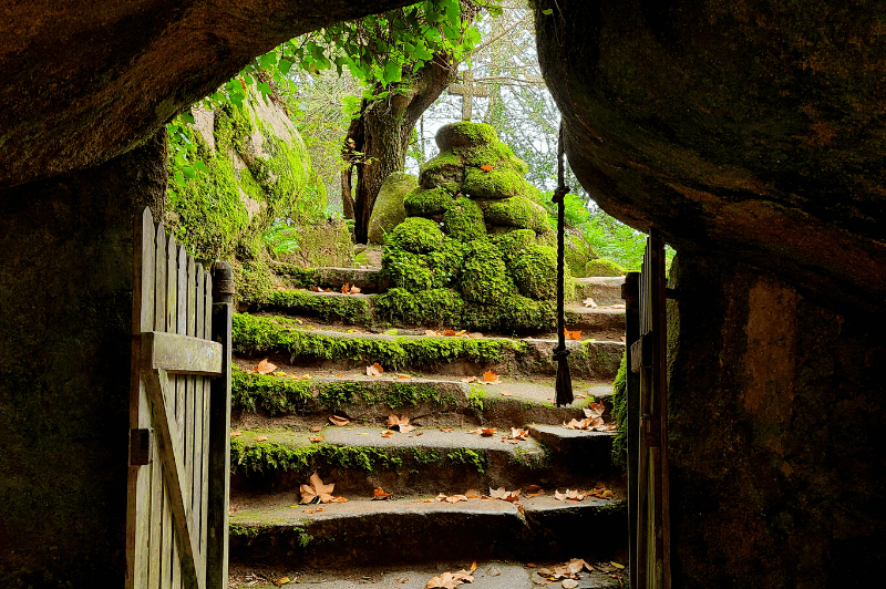 Boulder gate at the Convent of the Capuchos. Two large mossy boulders have fallen together to make a gate, with stairs leading up to a pile of stones with a crucifix on top.