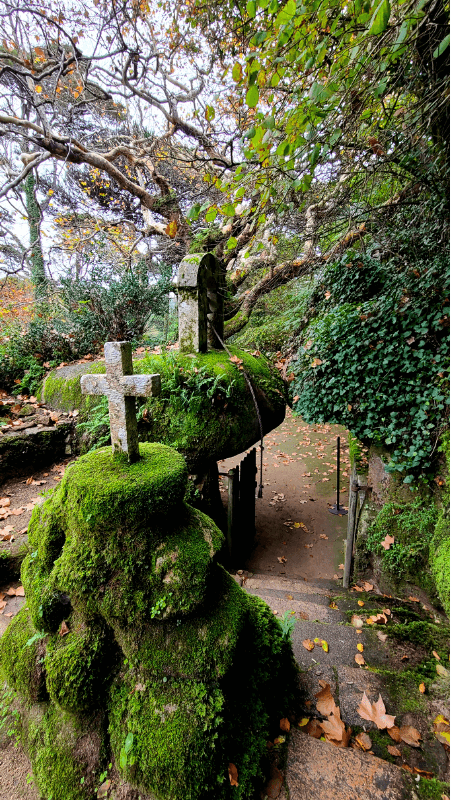 The courtyard of the bell at the Convent of the Capuchos features a cross on top a pile of mossy stones, and the bell over boulder gate in the background.