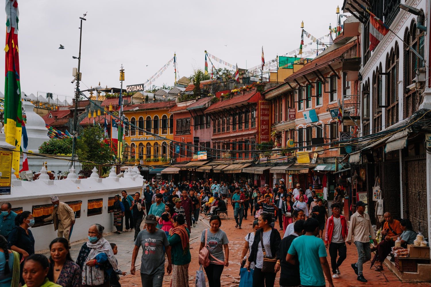 Locals walking around the Boudhanath Stupa kora with shops and cafes in the background.