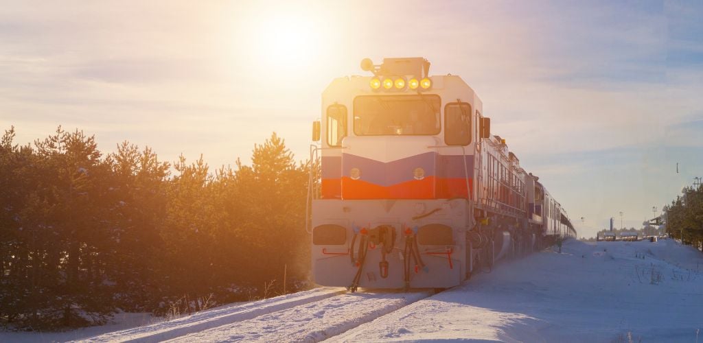 Is a train with snow on the tracks and trees on the side against the backdrop of a sunset. 
