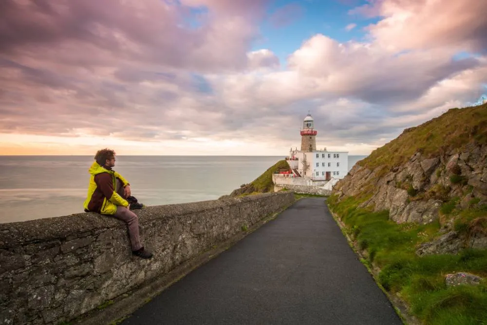 Man sitting on wall at sunset looking out to sea and lighthouse by Howth coastal path — Getty Images