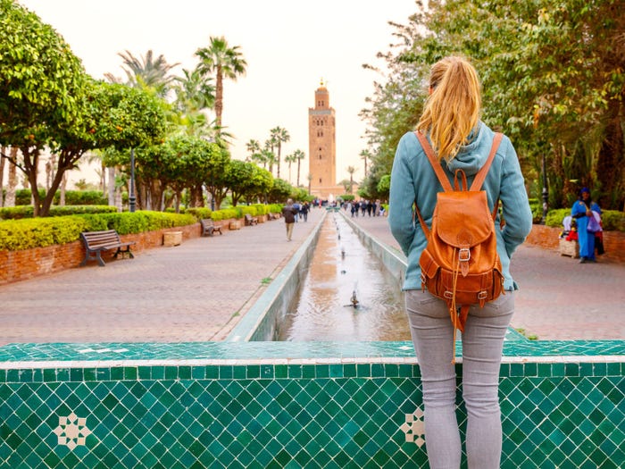 A woman, with an orange backpack, strawberry-blonde hair in a ponytail, and her back facing the camera, looks out at a mosque in Morocco.