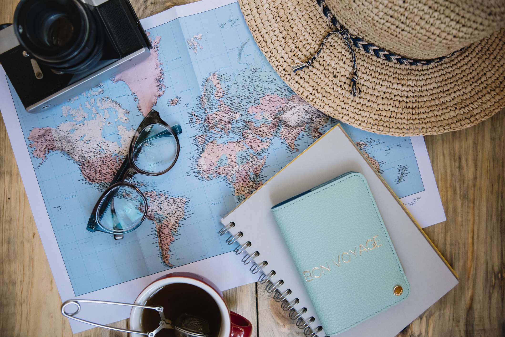 A map laid out on a table with a camera, notebook, coffee cup, hat and travel itinerary.