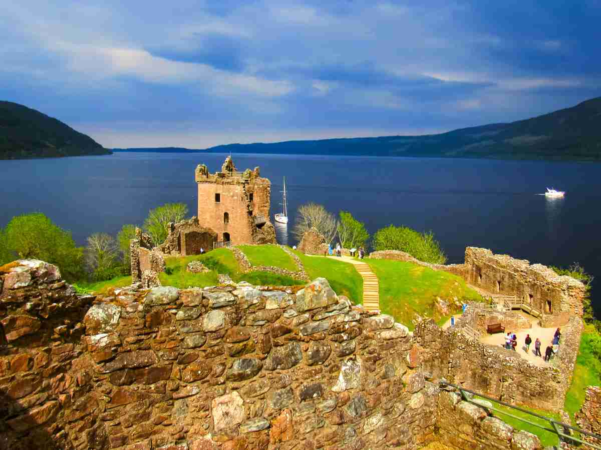 Urquhart Castle ruins near Drumnadrochit, with serene blue of Loch Ness as the backdrop. Tourists walking around.