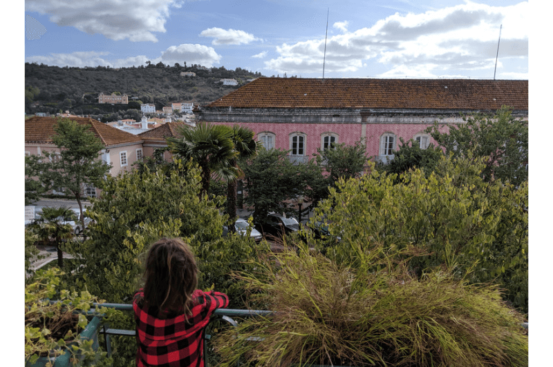 A small girl looks out over a railing onto a treed square. A pink tile building pokes through the trees on the far side.