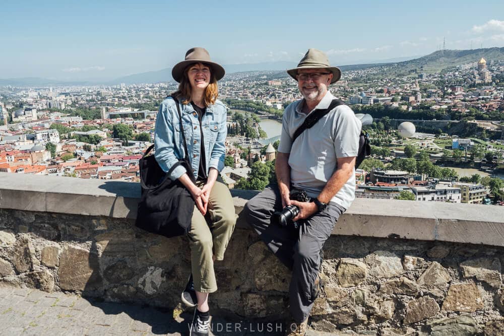 A woman and a man wearing hats sitting on a stone wall overlooking the city of Tbilisi, Georgia.