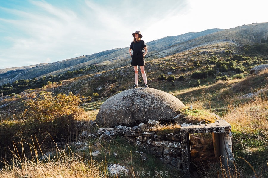 A woman dressed in a hat standing on top of a concrete bunker in a field in Albania.