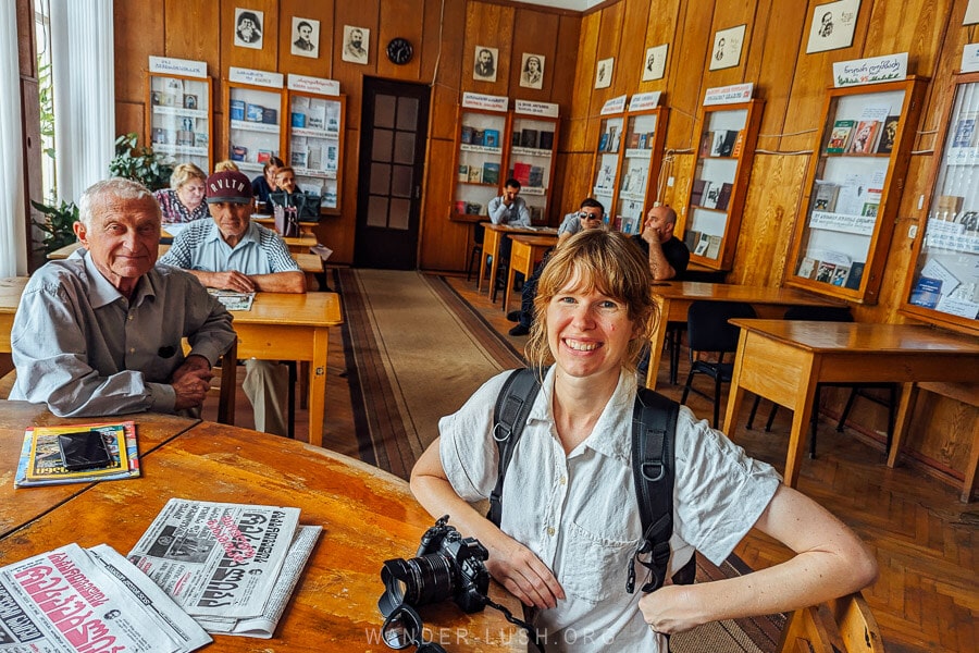 A woman sitting in a local library in Senaki, Georgia, with book shelves and patrons seated behind wooden reading desks.