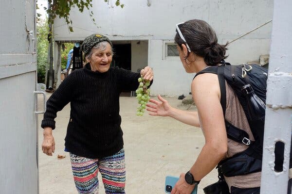 An older woman in a black sweater and colorful striped pants holds out a bunch of green grapes to a hiker wearing a sleeveless shirt, a black backpack and holding a smartphone in one hand. 