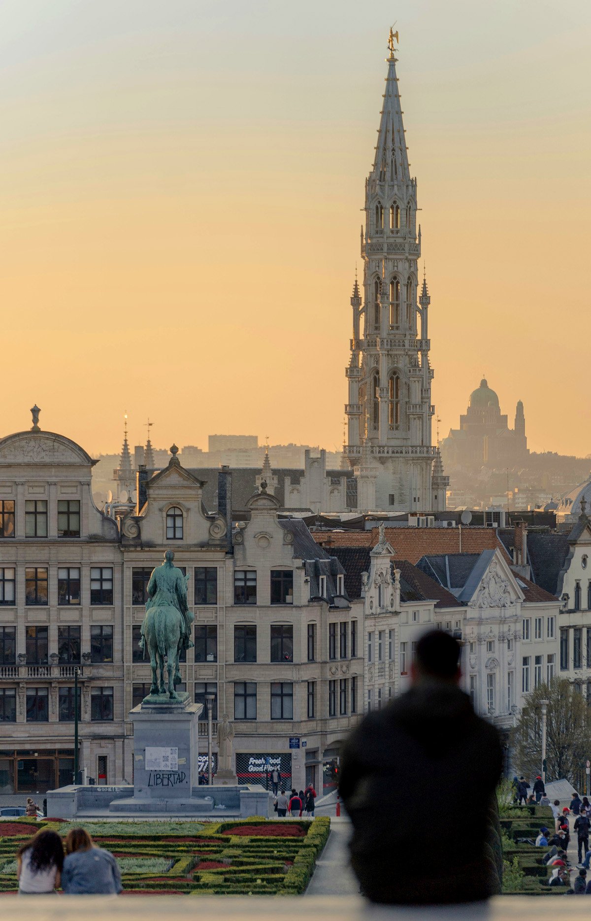 A man sits in a spot overlooking Brussels at sunset with a tall tower in the background. 