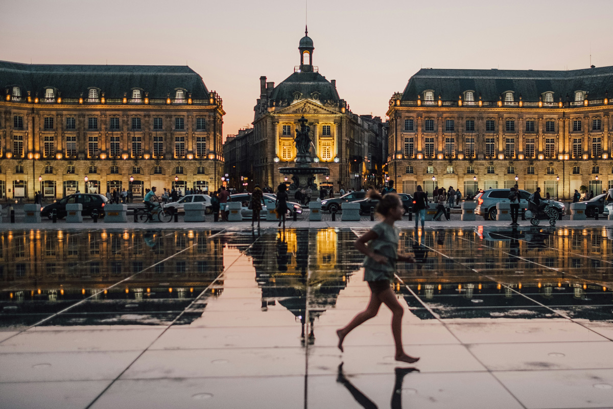 Buildings in Bordeaux are reflected in an enormous pool with a young girl running past in the foreground.